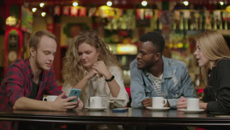 Portrait-of-cheerful-young-friends-looking-at-smart-phone-while-sitting-in-cafe.-Mixed-race-people-sitting-at-a-table-in-restaurant-using-mobile-phone